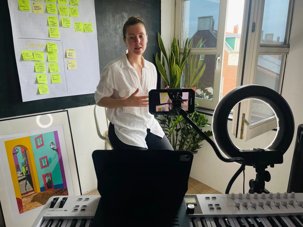 One of the crew in a white shirt talking to a phone on a tripod beside a ring light, a sticky-note planning board behind her and a music keyboard in front