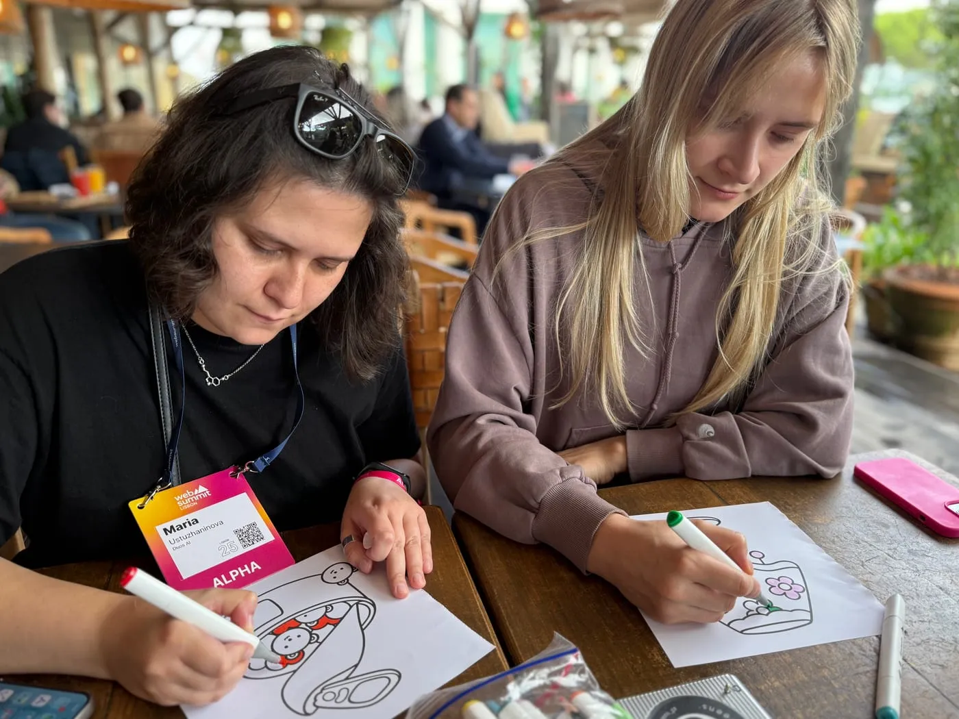 Two of the crew at a café table at Web Summit, badges on, colouring in pictures with felt-tip pens