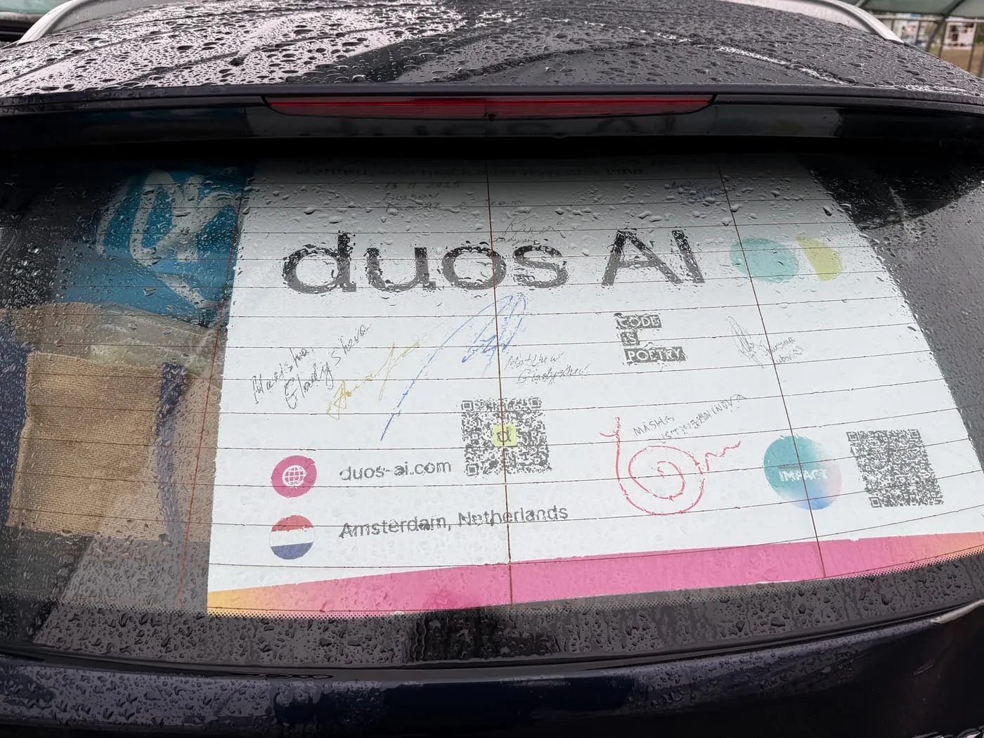 The Duos AI stand's banner in a car's rear window, covered in the crew's signatures, rain on the glass