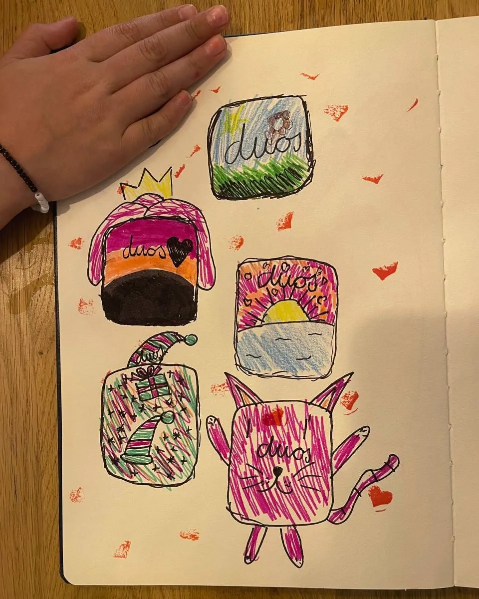 A small hand on a sketchbook page of five felt-tip Duos app icons: a sunny meadow, a crowned stripy one with a heart, a sunrise over the sea, a festive one with Christmas hats and a present, and a waving pink cat, with red hearts stamped all around