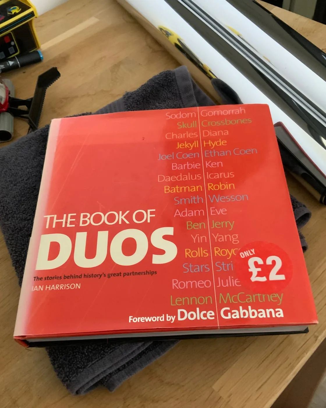 A red hardback on a desk, The Book of Duos by Ian Harrison, famous pairs listed down its cover and an "Only £2" sticker on it